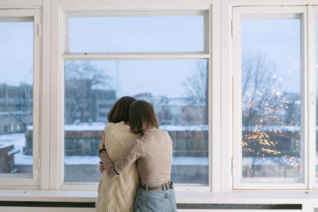 Two people hugging indoors near a window with a snowy, cityscape background.