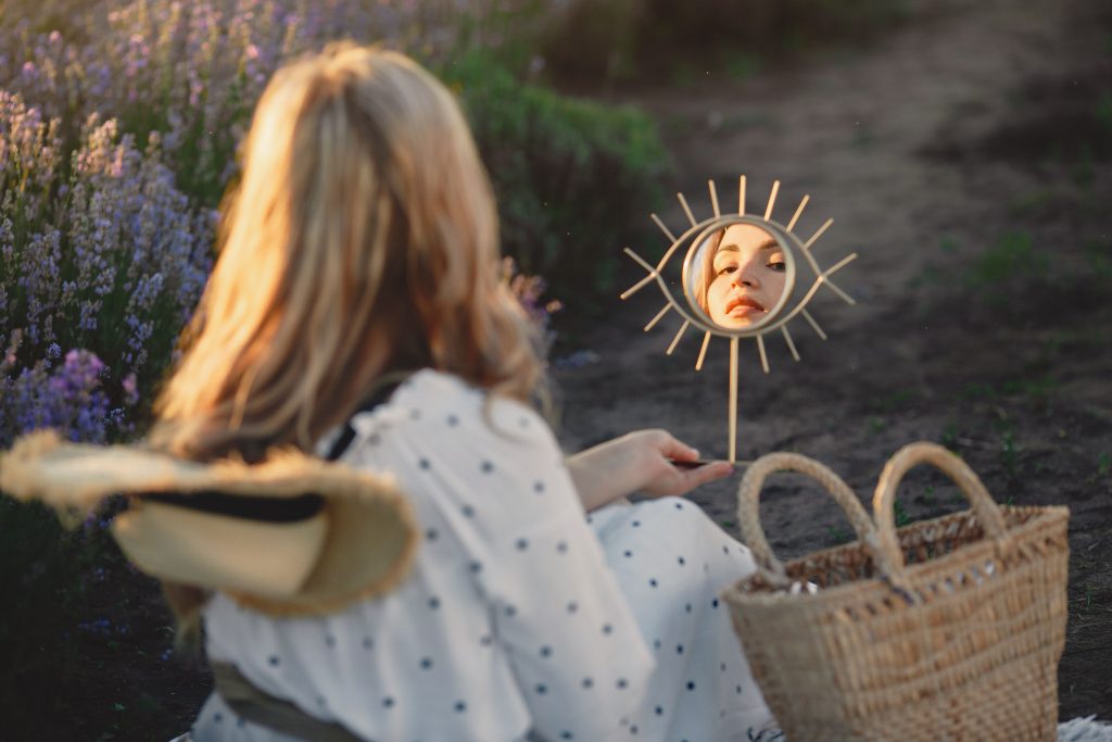 Blonde woman in field admiring reflection in a sun-shaped mirror, creating a whimsical scene.